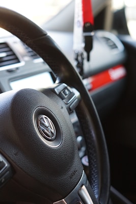 The image features a close-up view of a car's interior, specifically focusing on a Volkswagen steering wheel. Adjacent to the steering wheel, various controls and a portion of the dashboard are visible. A red and white keychain or accessory is hanging, and part of the car's air vents and infotainment system is present.