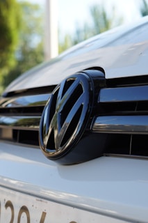 A close-up view of the front grille of a car, featuring the Volkswagen logo prominently displayed in the center. The logo is reflective, with black and chrome accents. The background is slightly blurred, suggesting the car is in an outdoor setting with greenery visible behind it.