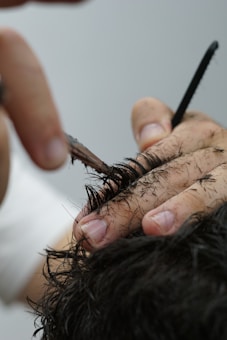 Hands are seen in the process of cutting hair with scissors. Hair clippings rest on the fingers of the person doing the haircut. The hair is dark, and the scene is closely focused on this grooming process.