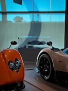 Several luxury sports cars are on display indoors, featuring vibrant colors and sleek designs. One car is prominently orange with distinct headlights, while another is white with black and red accents. A scale model of a car is showcased in a glass case at the center.
