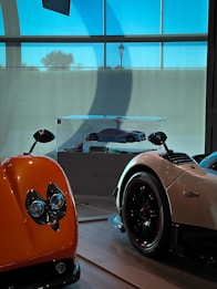 Several luxury sports cars are on display indoors, featuring vibrant colors and sleek designs. One car is prominently orange with distinct headlights, while another is white with black and red accents. A scale model of a car is showcased in a glass case at the center.
