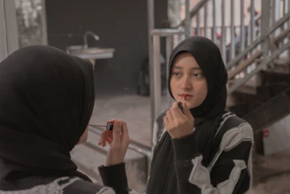 A person wearing a dark headscarf and a patterned sweater is applying makeup using a lipstick in front of a mirror. The background features a staircase and a sink against a muted setting, providing a casual and intimate atmosphere.