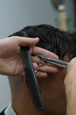 A close-up of a person getting a haircut. The hands of the barber are visible, one holding a comb and the other a straight razor. The dark, wet hair is being carefully trimmed, with small pieces of hair scattered on the barber's hand.
