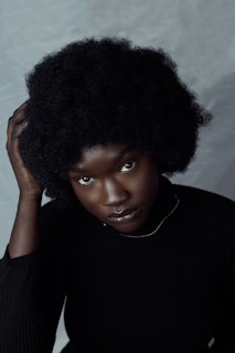 A person with a natural afro hairstyle is wearing a dark top against a textured gray background. Their expression is neutral and they are looking directly at the camera.