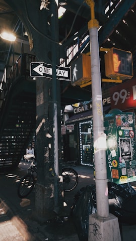 A gritty urban street scene featuring a one-way street sign, a pedestrian walk signal, and a cluttered utility pole covered with stickers. There is a staircase leading up to a raised platform or station. A bicycle is leaning against a column, and garbage bags are piled next to a pole. The area is dimly lit with beams of sunlight filtering through.