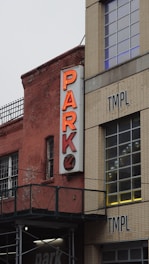 A neon sign with the word 'PARK' and an arrow pointing downwards is mounted on the side of a brick building. The building features large windows and a textured facade. Adjacent to it is another building labeled with the letters 'TMPL'. The structures appear industrial or urban, with visible scaffolding and railings.