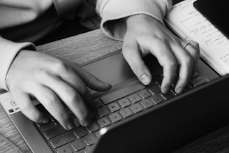 Hands typing on a laptop keyboard with a notepad beside, suggesting an active work or study environment. The focus is on the fingers pressing the keys, with a visible closed laptop and part of the notebook.