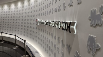 A curving wall decorated with small, white silhouette figures of various Pokémon. The wall is illuminated, highlighting the contours and creating a shadow effect. In the center, there is a three-dimensional sign with the text 'Pokémon Center TOKYO DX' featuring the Pokémon logo.