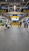 A railway platform scene with multiple people walking towards the camera. Overhead, there are digital displays showing train schedules in Japanese. The platform and its surroundings feature metal structural elements, and signage is prominently visible. There is a mixture of industrial and modern elements in the architecture.