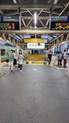 A railway platform scene with multiple people walking towards the camera. Overhead, there are digital displays showing train schedules in Japanese. The platform and its surroundings feature metal structural elements, and signage is prominently visible. There is a mixture of industrial and modern elements in the architecture.
