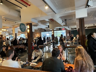 A bustling indoor market space with multiple people sitting and standing around tables enjoying food and drinks. The setting includes industrial-style ceilings with visible ducts and lighting fixtures, and exposed brick walls. There are signs with 'Time Out Market' branding visible. Natural light is coming through large windows in the background.