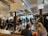 A bustling indoor market space with multiple people sitting and standing around tables enjoying food and drinks. The setting includes industrial-style ceilings with visible ducts and lighting fixtures, and exposed brick walls. There are signs with 'Time Out Market' branding visible. Natural light is coming through large windows in the background.
