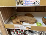 A shelf display contains olive wood serving and cutting boards stacked on top of each other. The shelf has a gold sign that advertises 'Cinnamon Bun Inspired Truffles' priced at $4.99. Below these boards, multicolored round felt items are visible, resembling coasters or trivets. Another label indicates a special 'Holiday Bazaar' and promotes the olive wood boards as 'gorgeous & long-lasting' for $12.99 each.