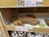 A shelf display contains olive wood serving and cutting boards stacked on top of each other. The shelf has a gold sign that advertises 'Cinnamon Bun Inspired Truffles' priced at $4.99. Below these boards, multicolored round felt items are visible, resembling coasters or trivets. Another label indicates a special 'Holiday Bazaar' and promotes the olive wood boards as 'gorgeous & long-lasting' for $12.99 each.