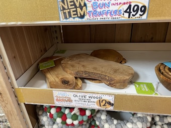 A shelf display contains olive wood serving and cutting boards stacked on top of each other. The shelf has a gold sign that advertises 'Cinnamon Bun Inspired Truffles' priced at $4.99. Below these boards, multicolored round felt items are visible, resembling coasters or trivets. Another label indicates a special 'Holiday Bazaar' and promotes the olive wood boards as 'gorgeous & long-lasting' for $12.99 each.