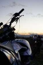 A motorcycle is prominently featured in the foreground with details such as a visible brand logo and the handlebars slightly silhouetted against a sunset. Another motorcycle is seen blurred in the background, suggesting an outdoor setting with a peaceful, open landscape.