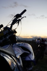 A motorcycle is prominently featured in the foreground with details such as a visible brand logo and the handlebars slightly silhouetted against a sunset. Another motorcycle is seen blurred in the background, suggesting an outdoor setting with a peaceful, open landscape.