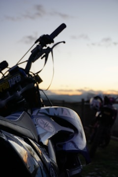 A motorcycle is prominently featured in the foreground with details such as a visible brand logo and the handlebars slightly silhouetted against a sunset. Another motorcycle is seen blurred in the background, suggesting an outdoor setting with a peaceful, open landscape.