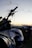 A motorcycle is prominently featured in the foreground with details such as a visible brand logo and the handlebars slightly silhouetted against a sunset. Another motorcycle is seen blurred in the background, suggesting an outdoor setting with a peaceful, open landscape.