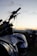 A motorcycle is prominently featured in the foreground with details such as a visible brand logo and the handlebars slightly silhouetted against a sunset. Another motorcycle is seen blurred in the background, suggesting an outdoor setting with a peaceful, open landscape.