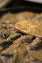 A close-up view of a creamy dish with visible chunks and a wooden spatula partially submerged in the mixture. The texture appears thick and smooth, with a glossy sheen reflecting light.