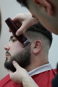 A person is receiving a haircut, with a focus on beard trimming. The barber is using electric clippers to shape the beard. The person is seated and draped in a red barber cape.