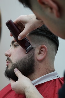 A person is receiving a haircut, with a focus on beard trimming. The barber is using electric clippers to shape the beard. The person is seated and draped in a red barber cape.