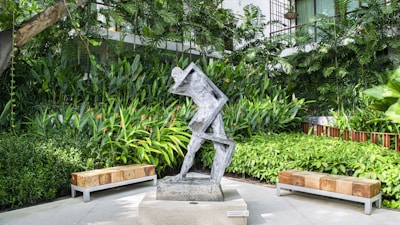 A modern abstract sculpture made of metal is placed on a stone pedestal in a garden setting. The sculpture features angular, geometric shapes that appear to represent a human figure in motion. It is surrounded by lush, green tropical plants and trees, creating a serene and peaceful atmosphere. Two wooden benches are positioned symmetrically on either side of the sculpture.