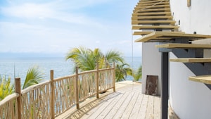 A wooden deck overlooking the ocean, with palm trees in the background and a modern staircase on the right side. The sky is clear with a few clouds, creating a serene and tropical setting.