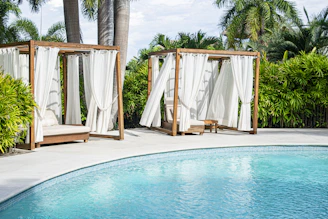 Two wooden cabanas with white curtains are positioned next to a clear blue swimming pool, surrounded by lush green palm trees and bushes.