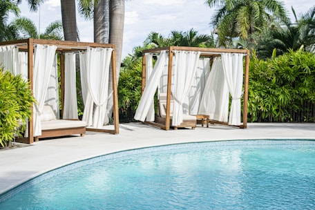 Two wooden cabanas with white curtains are positioned next to a clear blue swimming pool, surrounded by lush green palm trees and bushes.