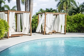 Two wooden cabanas with white curtains are positioned next to a clear blue swimming pool, surrounded by lush green palm trees and bushes.