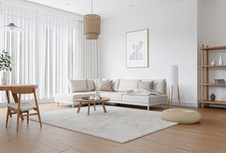 A modern living room with a minimalist design featuring a large beige sectional sofa adorned with light colored pillows. A round wooden coffee table sits on a fluffy light beige rug. A wooden dining table with two chairs is in the corner. Natural light fills the room through tall white sheer curtains. The walls are white, with a framed abstract art piece hanging above the sofa. Other elements include a wooden floor lamp, a woven floor pouf, a hanging rattan light fixture, and a wooden bookshelf with decor items.
