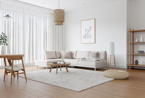 A modern living room with a minimalist design featuring a large beige sectional sofa adorned with light colored pillows. A round wooden coffee table sits on a fluffy light beige rug. A wooden dining table with two chairs is in the corner. Natural light fills the room through tall white sheer curtains. The walls are white, with a framed abstract art piece hanging above the sofa. Other elements include a wooden floor lamp, a woven floor pouf, a hanging rattan light fixture, and a wooden bookshelf with decor items.