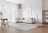 A modern living room with a minimalist design featuring a large beige sectional sofa adorned with light colored pillows. A round wooden coffee table sits on a fluffy light beige rug. A wooden dining table with two chairs is in the corner. Natural light fills the room through tall white sheer curtains. The walls are white, with a framed abstract art piece hanging above the sofa. Other elements include a wooden floor lamp, a woven floor pouf, a hanging rattan light fixture, and a wooden bookshelf with decor items.