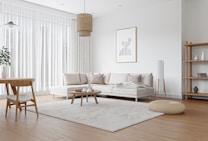 A modern living room with a minimalist design featuring a large beige sectional sofa adorned with light colored pillows. A round wooden coffee table sits on a fluffy light beige rug. A wooden dining table with two chairs is in the corner. Natural light fills the room through tall white sheer curtains. The walls are white, with a framed abstract art piece hanging above the sofa. Other elements include a wooden floor lamp, a woven floor pouf, a hanging rattan light fixture, and a wooden bookshelf with decor items.