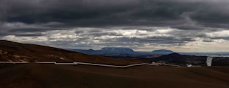 A vast, barren landscape stretches across the horizon. The sky is filled with thick, dark clouds, creating a dramatic atmosphere. A long pipeline snakes its way through the rugged terrain, with mountains visible in the background. In the distance, there is a plume of steam rising from the ground.