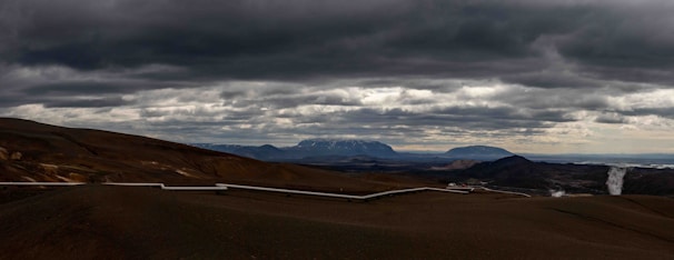 A vast, barren landscape stretches across the horizon. The sky is filled with thick, dark clouds, creating a dramatic atmosphere. A long pipeline snakes its way through the rugged terrain, with mountains visible in the background. In the distance, there is a plume of steam rising from the ground.