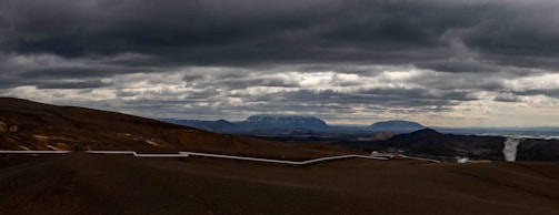 A vast, barren landscape stretches across the horizon. The sky is filled with thick, dark clouds, creating a dramatic atmosphere. A long pipeline snakes its way through the rugged terrain, with mountains visible in the background. In the distance, there is a plume of steam rising from the ground.