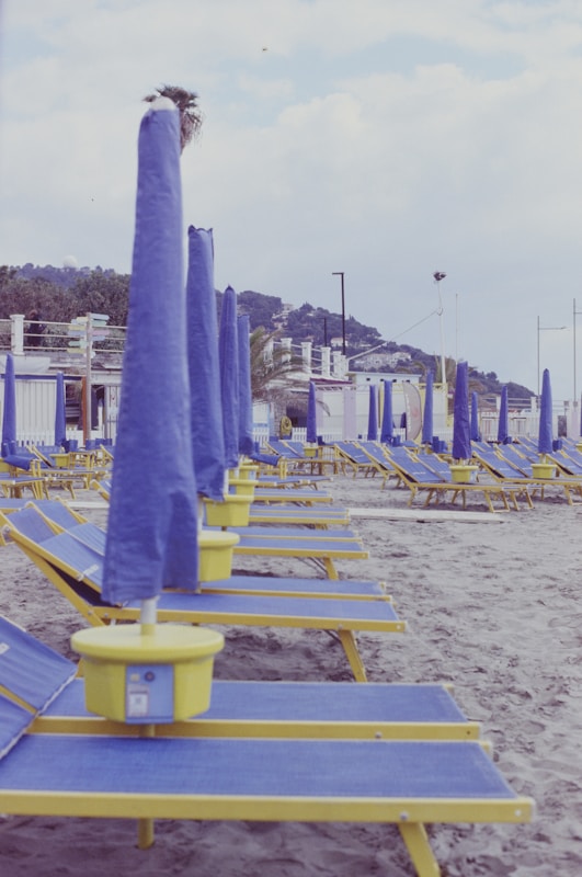 Rows of empty beach loungers with closed blue umbrellas stand on a sandy beach. The loungers are yellow with blue cushions, and there are small yellow tables attached to each one. In the background, there are several beach huts and some hills with greenery and buildings.