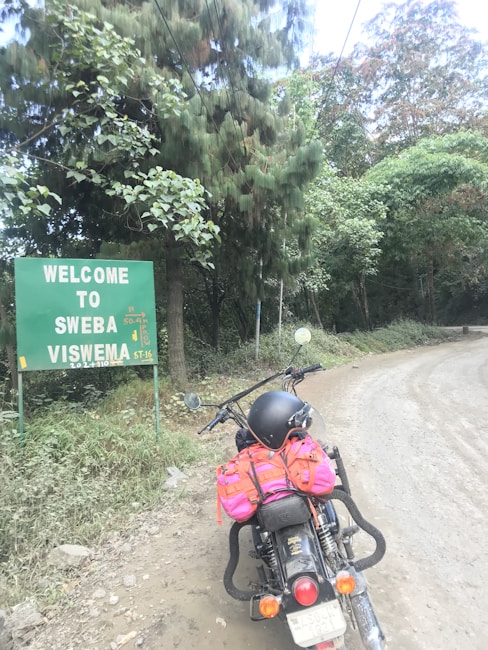 A motorcycle is parked on a dirt road near a green sign that reads 'Welcome to Swebab Viswema'. The bike has a pink and orange backpack and a black helmet resting on the seat. Tall trees with dense foliage surround the area.