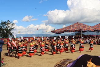 A group of people dressed in traditional, colorful attire are performing a cultural dance outdoors. The scene is lively, with vibrant fabrics and intricate patterns. A large, decorative tent canopy is set up in the background with an audience watching the performance. Flags are positioned around the area, and trees are visible in the distance under a partly cloudy sky.