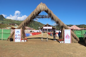 A large outdoor event setup with a grassy archway entrance leads to a stage area. Rows of red and blue chairs are arranged neatly in front of the stage. The surrounding area includes green hills and a clear blue sky. Welcome banners featuring photographs are placed at the entrance, and colorful tents are seen on the sides.