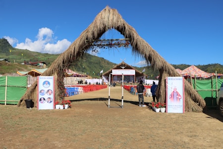 A large outdoor event setup with a grassy archway entrance leads to a stage area. Rows of red and blue chairs are arranged neatly in front of the stage. The surrounding area includes green hills and a clear blue sky. Welcome banners featuring photographs are placed at the entrance, and colorful tents are seen on the sides.