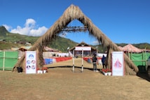 A large outdoor event setup with a grassy archway entrance leads to a stage area. Rows of red and blue chairs are arranged neatly in front of the stage. The surrounding area includes green hills and a clear blue sky. Welcome banners featuring photographs are placed at the entrance, and colorful tents are seen on the sides.