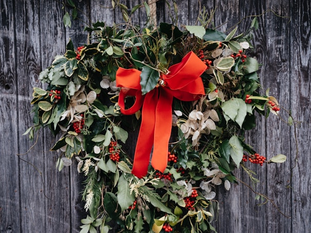 A heart-shaped wreath made of various green leaves, berries, and adorned with a large, bright red ribbon is hanging against a weathered wooden background. The foliage is lush and includes both glossy and matte textures, while clusters of red berries add vibrant pops of color.