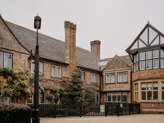A rustic brick house with multiple chimneys, surrounded by a wooden fence and some greenery. The building features traditional architectural elements such as a gabled roof and grid-patterned windows. A lantern pole stands in the foreground.