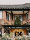 A traditional brick building with white-framed windows is adorned with autumnal ivy and greenery. At the entrance, an arched sign reads 'Hotel Entrance,' and a warm lantern light hangs above, creating an inviting atmosphere.