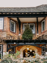 A traditional brick building with white-framed windows is adorned with autumnal ivy and greenery. At the entrance, an arched sign reads 'Hotel Entrance,' and a warm lantern light hangs above, creating an inviting atmosphere.