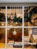 A cozy interior view of a cafe adorned with holiday decorations, including evergreen garlands with pinecones and hanging glass ornaments. A small gingerbread house sits on a wooden counter alongside a stack of books and games. Warm lighting creates a welcoming ambiance.